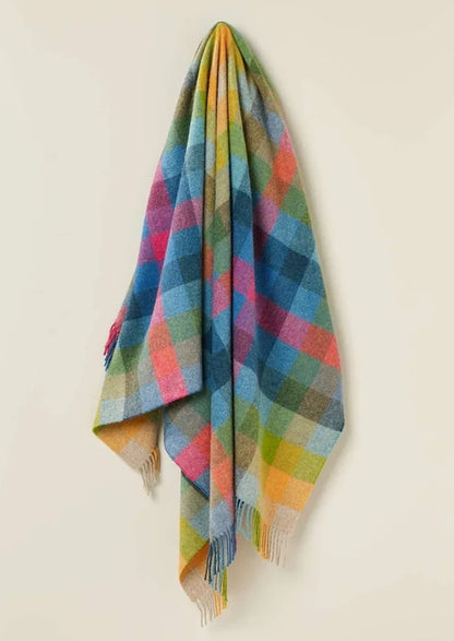 A multicolor plaid wool throw blanket with fringe, hanging against a plain off-white background.