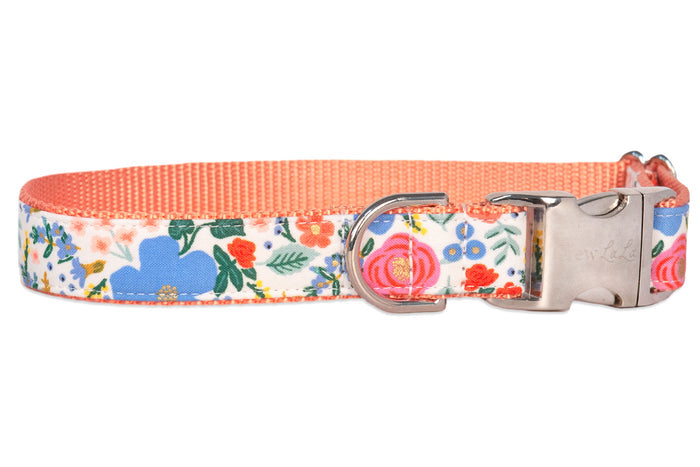 A fabric dog collar with a floral pattern featuring blue, pink, orange, and green elements on a white background. The collar has a peachy orange inner webbing, a silver metal D-ring, and a silver side-release buckle.