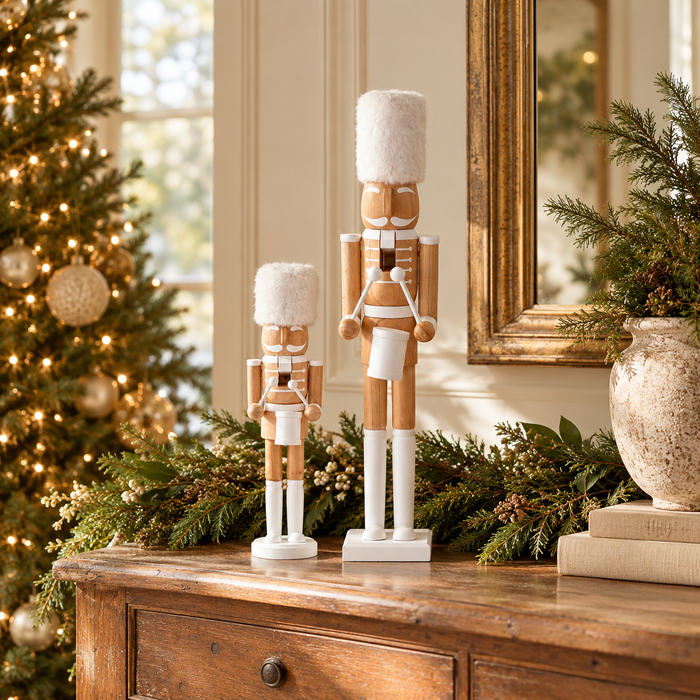 Two wooden nutcracker figurines with white fur-like hats and coats are displayed on a wooden console table in a decorated holiday room, with a Christmas tree and mirror in the background.